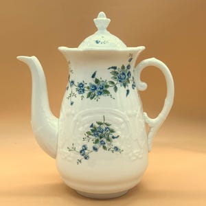 Pode incluir: Bule de cerâmica branco com tampa e bico curvo. O bule é decorado com padrões florais azuis e tem uma alça. O bule é de estilo vintage e provavelmente é usado para servir chá.