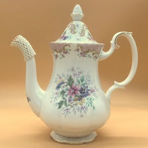 Può includere: Caffettiera in porcellana bianca con un motivo floreale rosa, blu e viola. La caffettiera ha un manico curvo e un coperchio con un pomello decorativo. Il beccuccio è protetto da una rete bianca.