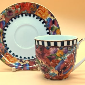 Puede incluir: Un juego de taza y platillo con motivos florales. La taza y el platillo tienen una base blanca con un colorido diseño floral y de mariposas. La taza tiene un borde a rayas blanco y negro y un asa dorada.