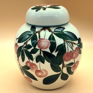 Może przedstawiać: Biały ceramiczny słoik z pokrywką, ozdobiony ręcznie malowanym wzorem gałęzi wiśni. Wiśnie są w kolorze miękkiej czerwieni, a liście są zielone. Słoik ma okrągły kształt.