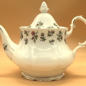 Może przedstawiać: Biały porcelanowy imbryk z kwiatowym wzorem z różowych i niebieskich kwiatów. Imbryk ma zakrzywiony uchwyt, dzióbek i pokrywkę z guzikiem. Podstawa i obręcz są wykończone złotem. Klasyczny element do serwowania herbaty.