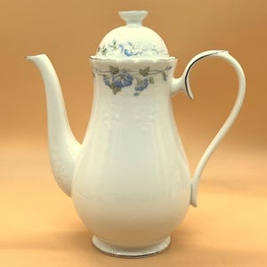 Pode incluir: Bule de café de porcelana branca com um design floral com flores azuis e folhas verdes. A bule tem uma tampa com uma borda festonada e uma borda prateada.