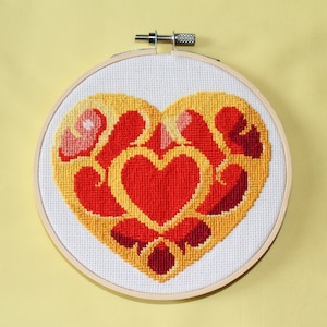 May include: A cross-stitch embroidery hoop with a red and yellow heart design. The heart is surrounded by a yellow border and has a smaller red heart in the center.