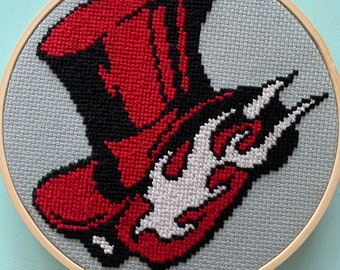 Może przedstawiać: Haftowany krzyżykiem czerwony i czarny wzór logo Persona 5, przedstawiający cylinder z motywem płomienia, w drewnianym obręczy do haftu.
