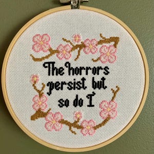 以下が含まれることがあります： 木製のフレームに入ったクロスステッチ刺繍。デザインは、黒で「The horrors persist but so do I」というテキストと、ピンクの桜と茶色の枝で構成されています。