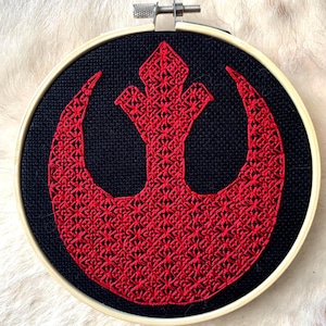 May include: A red embroidered Rebel Alliance symbol on a black fabric background in a wooden embroidery hoop.