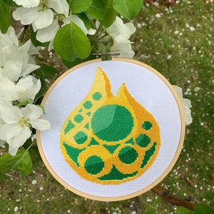 以下が含まれることがあります： 黄色と緑色の炎のようなデザインのクロスステッチ刺繍枠。 デザインは白い布にあり、木の枠で保持されています。 背景には白い花と緑の葉があります。