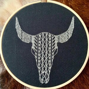 May include: A black fabric hoop features a white embroidered bull skull with intricate geometric patterns. The embroidery is detailed, with a mix of lines and shapes. The hoop is set in a wooden frame, creating a rustic, handcrafted aesthetic.