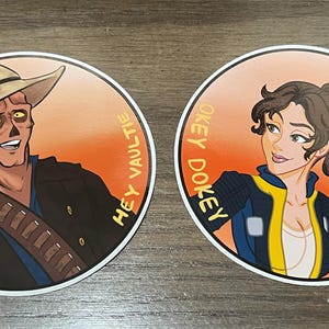 May include: Two round stickers with orange backgrounds and white borders. The left sticker features a cartoon man with a brown hat and a black jacket. The text "Hey Vaultie" is written vertically on the right side of the sticker. The right sticker features a cartoon woman with brown hair wearing a blue jacket with a yellow stripe. The text "Okey Dokey" is written vertically on the left side of the sticker.