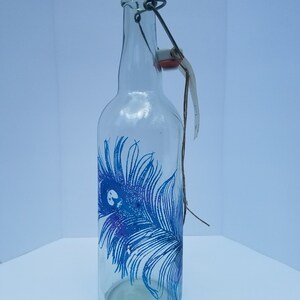 Puede incluir: Una botella de vidrio transparente con un diseño de pluma de pavo real azul. La botella tiene un cierre de alambre con un tapón rojo.