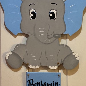 May include: Wooden baby elephant wall hanging with blue ears and a blue nameplate that reads "Benjamin". The elephant is gray with white accents. A space below the nameplate is available for baby's birth details.