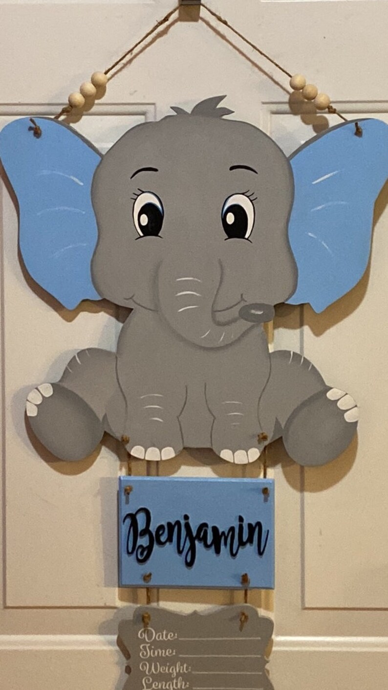 May include: Wooden baby elephant wall hanging with blue ears and a blue nameplate that reads "Benjamin". The elephant is gray with white accents. A space below the nameplate is available for baby's birth details.