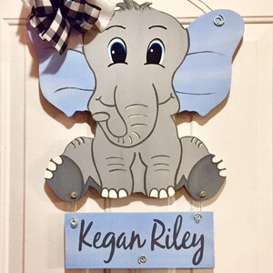 May include: A wooden elephant-shaped door hanger with blue ears and a matching sign that reads "Kegan Riley." The elephant has a gray body and a black and white checkered bow. A smaller sign below has space for birth details.
