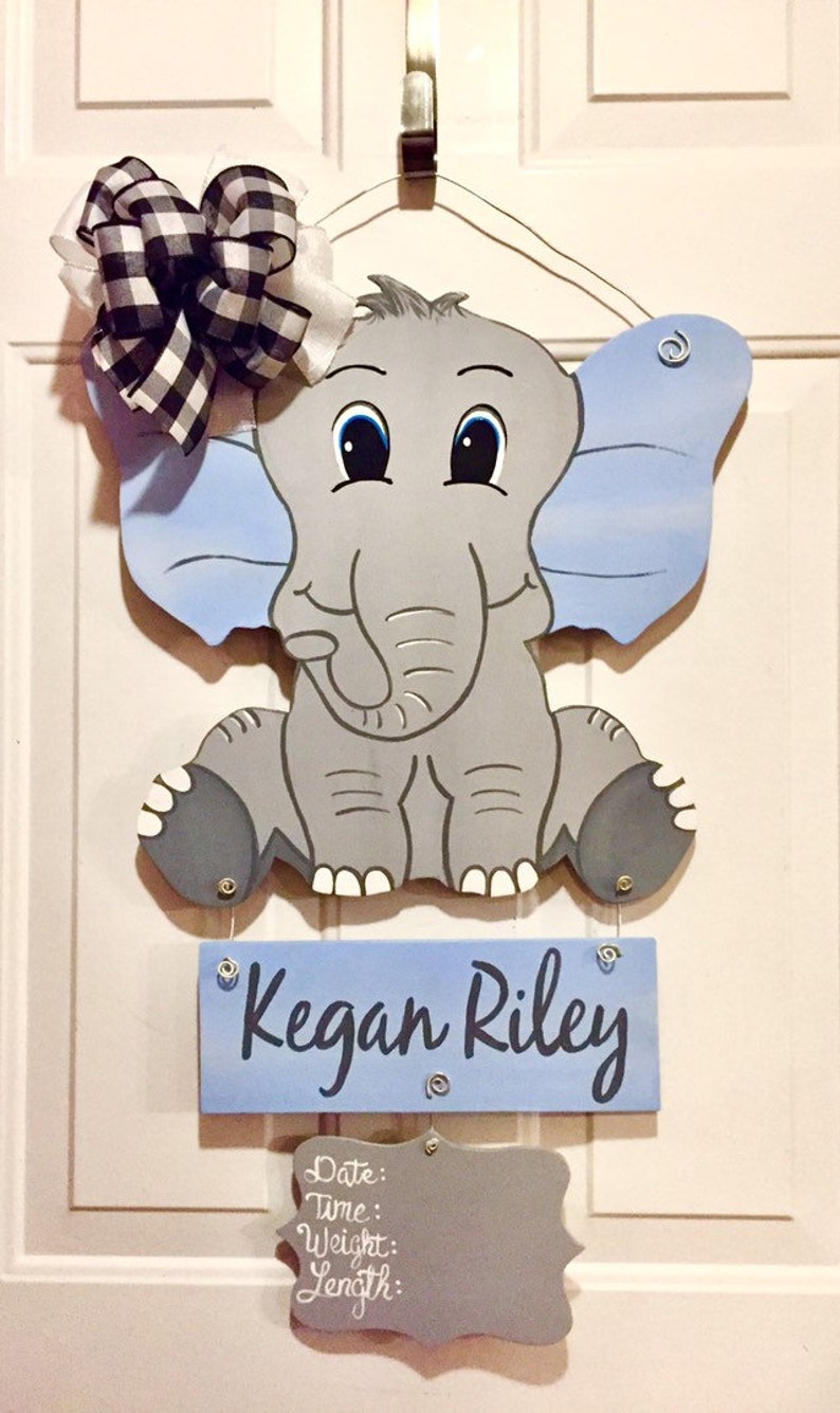 May include: A wooden elephant-shaped door hanger with blue ears and a matching sign that reads "Kegan Riley." The elephant has a gray body and a black and white checkered bow. A smaller sign below has space for birth details.