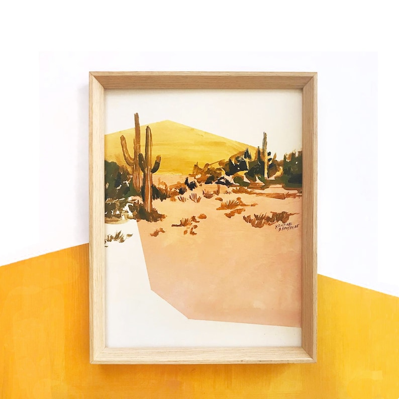 May include: A watercolor painting of a desert landscape with tall cacti and a yellow and orange sky. The painting is framed in a light wood frame.