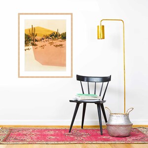 May include: A framed print of a desert landscape with two cacti and a yellow hill in the background. The print is hanging on a white wall above a black chair and a silver woven basket.