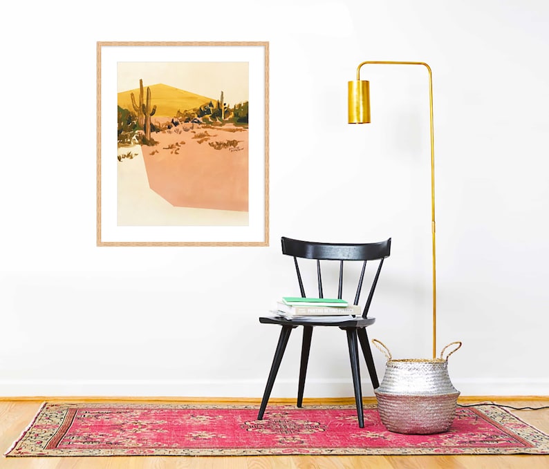 May include: A framed print of a desert landscape with two cacti and a yellow hill in the background. The print is hanging on a white wall above a black chair and a silver woven basket.