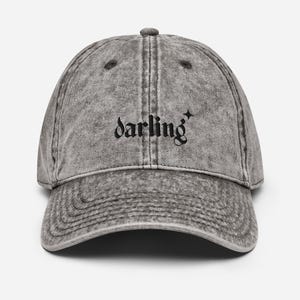 May include: A gray, vintage-style baseball cap with the word "darling" in black script across the front. A small star graphic is next to the word. The cap has a curved brim and a fabric strap closure.