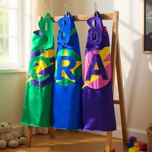 May include: Three colorful superhero capes hanging on a wooden rack. The capes are green, blue, and purple, each with a large letter and a lightning bolt design. A teddy bear and building blocks are in the foreground.