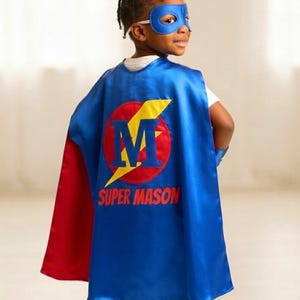 May include: A blue superhero cape with a red interior lining, featuring a circular emblem with a yellow lightning bolt and the letter M in blue. Below the emblem, the text SUPER MASON is printed in red. A matching blue eye mask is worn.