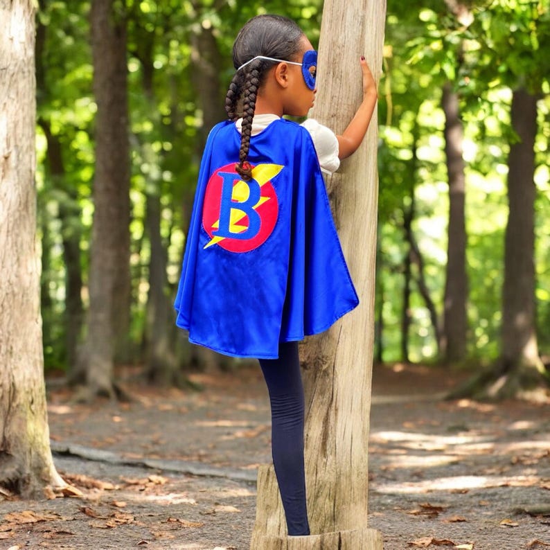Peut inclure: Un enfant portant une cape de super-h&eacute;ros bleue avec un cercle rouge et un &eacute;clair jaune et la lettre "B" au centre. L'enfant porte un masque pour les yeux bleu et a le dos tourn&eacute; vers la cam&eacute;ra.