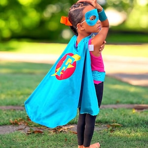 May include: A child wearing a blue superhero cape with a red circle and a yellow lightning bolt and the letter "F" on it. The child is also wearing a blue eye mask and a pink shirt.