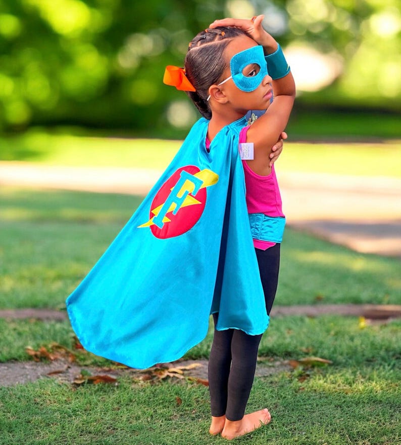 May include: A child wearing a blue superhero cape with a red circle and a yellow lightning bolt and the letter "F" on it. The child is also wearing a blue eye mask and a pink shirt.