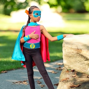 May include: A young person dressed as a superhero wearing a blue and red cape, a pink tank top, black leggings, a blue belt with a yellow "F" on it, and a blue mask. The person is standing next to a large rock.
