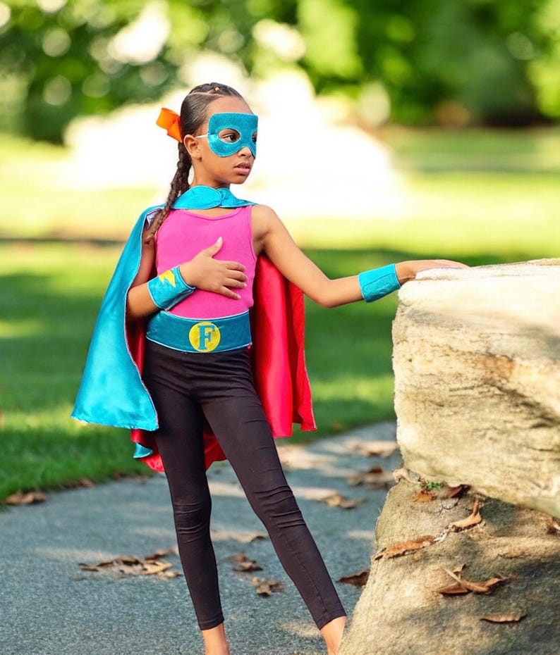 May include: A young person dressed as a superhero wearing a blue and red cape, a pink tank top, black leggings, a blue belt with a yellow "F" on it, and a blue mask. The person is standing next to a large rock.