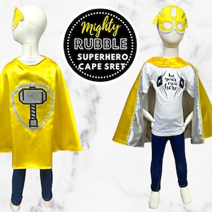 K&ouml;nnte beinhalten: Gelber Superhelden-Umhang mit einem silbernen Hammer-Design auf der R&uuml;ckseite. Der Umhang wird von einer Kinder-Schaufensterpuppe getragen. Das Kind tr&auml;gt auch ein wei&szlig;es T-Shirt mit dem Text "Sei dein eigener Held" in Schwarz.