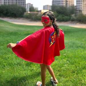 May include: A red superhero cape with a matching mask. The cape features a circular emblem with a lightning bolt and the initials "JW". The person is standing on a grassy field.