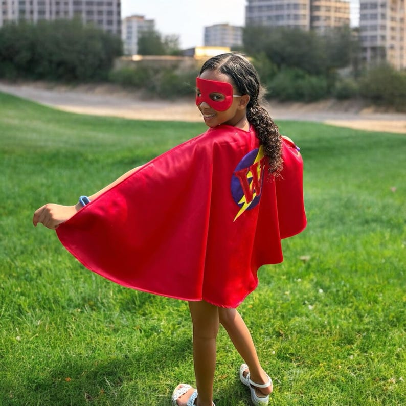 May include: A red superhero cape with a matching mask. The cape features a circular emblem with a lightning bolt and the initials "JW". The person is standing on a grassy field.