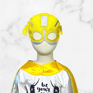 K&ouml;nnte beinhalten: Eine gelbe und silberne Superheldenmaske mit dem Text "be your own hero" auf einem wei&szlig;en T-Shirt mit einem gelben Umhang.