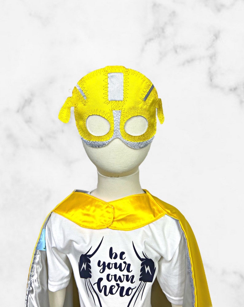 K&ouml;nnte beinhalten: Eine gelbe und silberne Superheldenmaske mit dem Text "be your own hero" auf einem wei&szlig;en T-Shirt mit einem gelben Umhang.