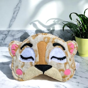 May include: A plush sleep mask shaped like a tiger face with tan and white fur, black embroidered closed eyes, a black nose, and pink accents on the ears and cheeks. The mask features a soft, fuzzy texture and includes an elastic strap for wear.