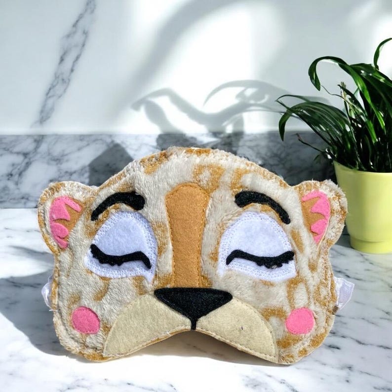 May include: A plush sleep mask shaped like a tiger face with tan and white fur, black embroidered closed eyes, a black nose, and pink accents on the ears and cheeks. The mask features a soft, fuzzy texture and includes an elastic strap for wear.