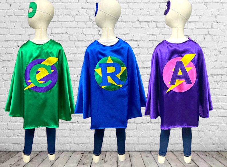Peut inclure: Trois capes de super-h&eacute;ros de couleurs diff&eacute;rentes, personnalis&eacute;es avec une lettre et un &eacute;clair. Les capes sont vertes, bleues et violettes.