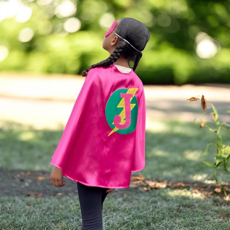 May include: A pink superhero cape with a green circle and a yellow lightning bolt. The letter "J" is in the centre of the circle. The cape is worn by a child with braided hair.