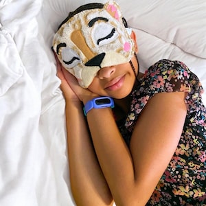 May include: A plush sleep mask shaped like a cat face with embroidered closed eyes, a black nose, and pink accents. The mask features a textured, cream-colored fabric with tan patches. A light blue digital watch is worn on the wrist.