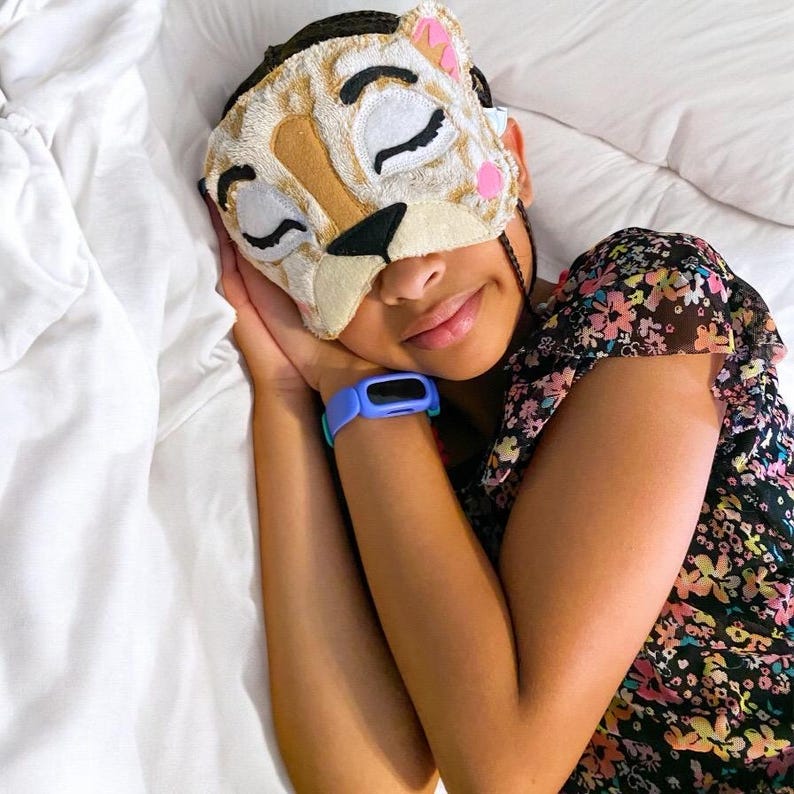 May include: A plush sleep mask shaped like a cat face with embroidered closed eyes, a black nose, and pink accents. The mask features a textured, cream-colored fabric with tan patches. A light blue digital watch is worn on the wrist.