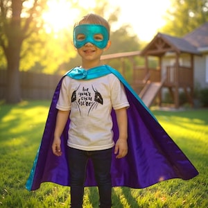 May include: A young child wearing a teal superhero mask and cape. The cape is teal on the top and purple on the bottom. The child is wearing a white t-shirt with the words "Be your own hero" printed on it.
