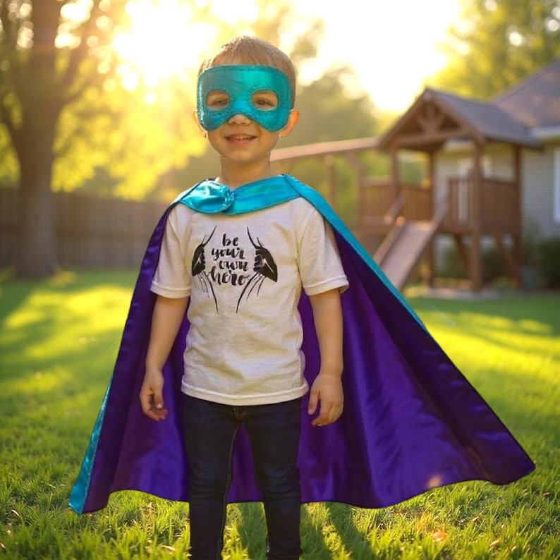 May include: A young child wearing a teal superhero mask and cape. The cape is teal on the top and purple on the bottom. The child is wearing a white t-shirt with the words "Be your own hero" printed on it.