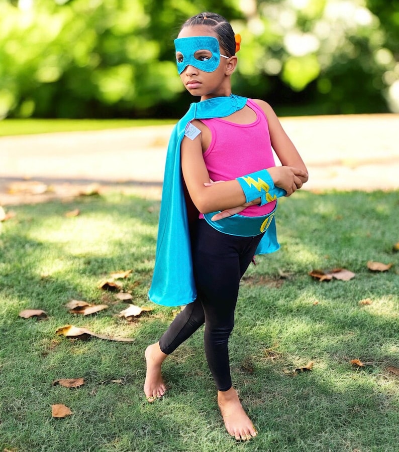 May include: A child is wearing a teal superhero costume. The costume includes a teal cape, a pink tank top, black leggings, and a teal eye mask.  The child also has on yellow and teal arm bands. The costume is accessorized with a teal belt.