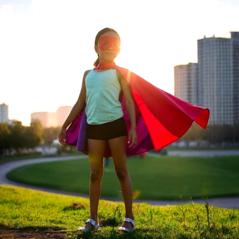 May include: A young person stands in a park, wearing a red superhero cape and mask. The cape is red on the outside and purple on the inside. They are wearing a light blue tank top and black shorts. The background includes green grass, trees, and buildings.
