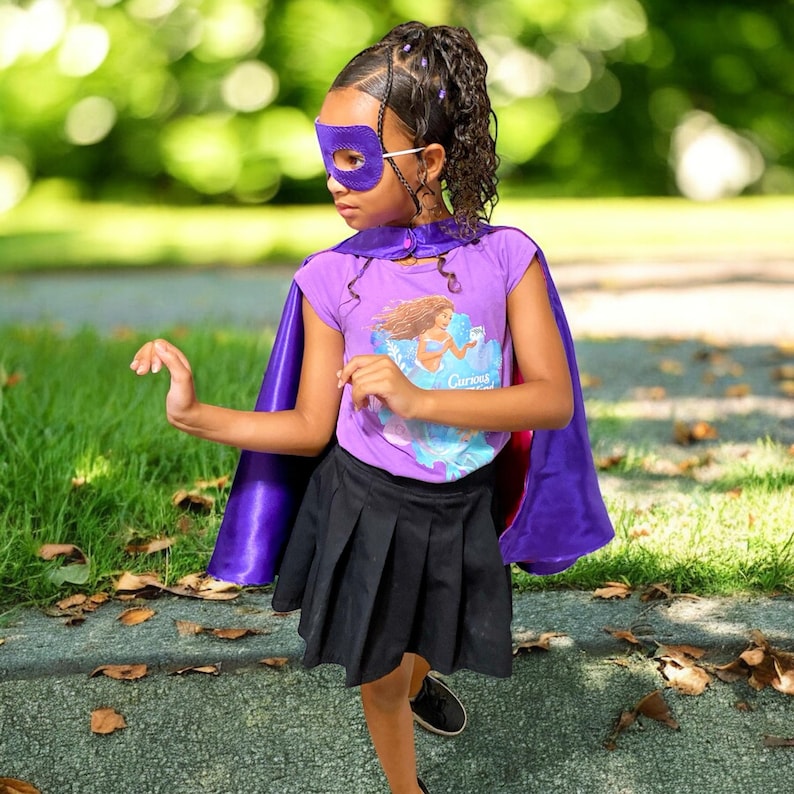 May include: A young person in a purple superhero costume, including a mask, cape, and t-shirt with a mermaid graphic. The outfit is paired with a black pleated skirt and sneakers. The background is a blurred outdoor scene.