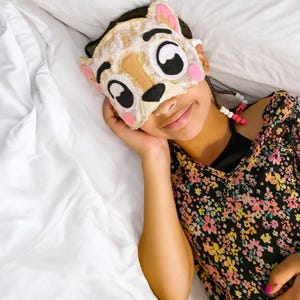 May include: A sleep mask shaped like a cat face with beige, white, and black felt details, including large eyes, a nose, and pink cheeks. The mask features small ears at the top and is secured with a strap adorned with white and pink beads.