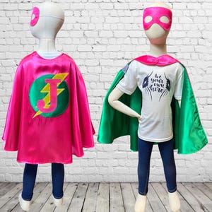 May include: A child wearing a pink and green superhero cape with a lightning bolt and the letter J on the back. The cape is made of satin and has a satin trim. The child is also wearing a white t-shirt with the text "Be your own hero" on the front. The child is wearing a pink mask.