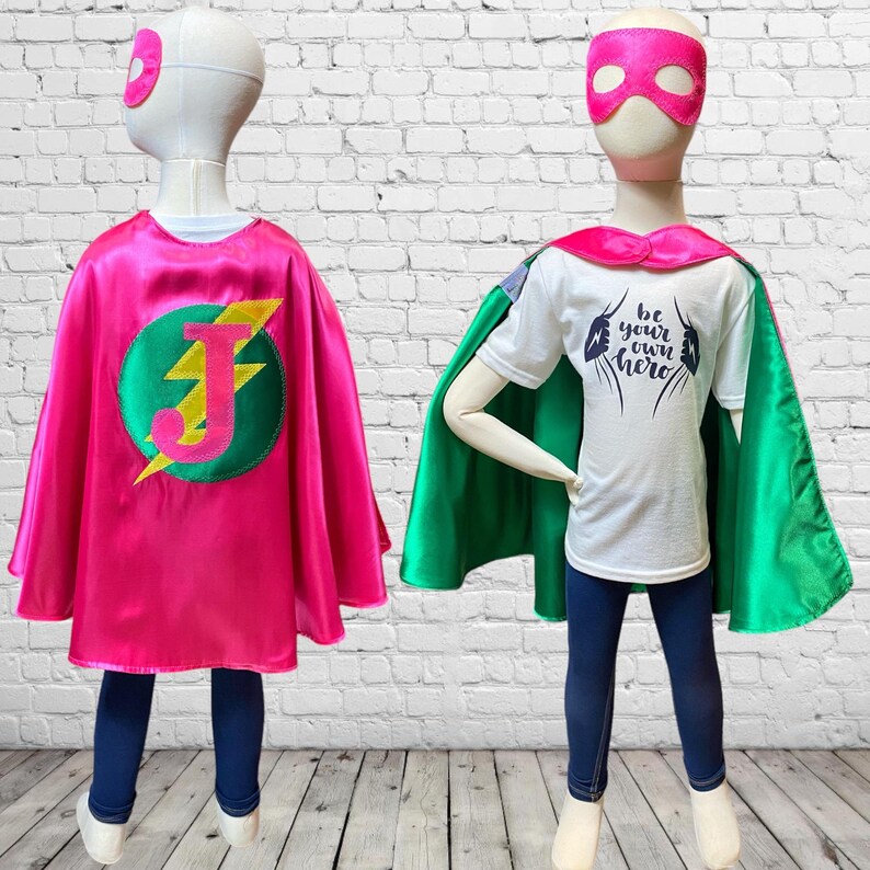 May include: A child wearing a pink and green superhero cape with a lightning bolt and the letter J on the back. The cape is made of satin and has a satin trim. The child is also wearing a white t-shirt with the text "Be your own hero" on the front. The child is wearing a pink mask.