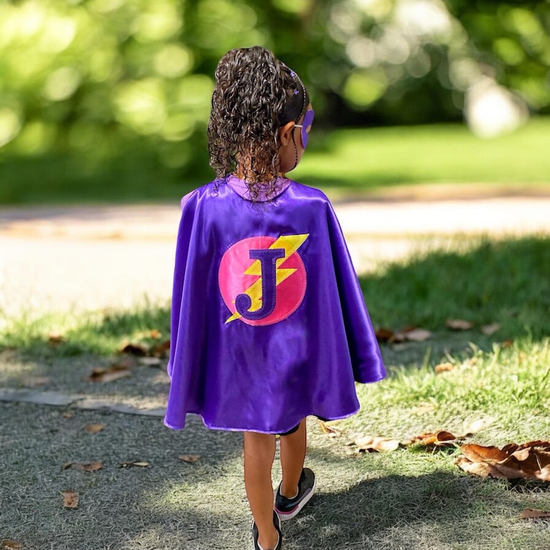 May include: A purple superhero cape with a pink circle, yellow lightning bolt, and the letter "J". The cape is worn by a person with a matching mask and headband. The person is walking on a paved path.
