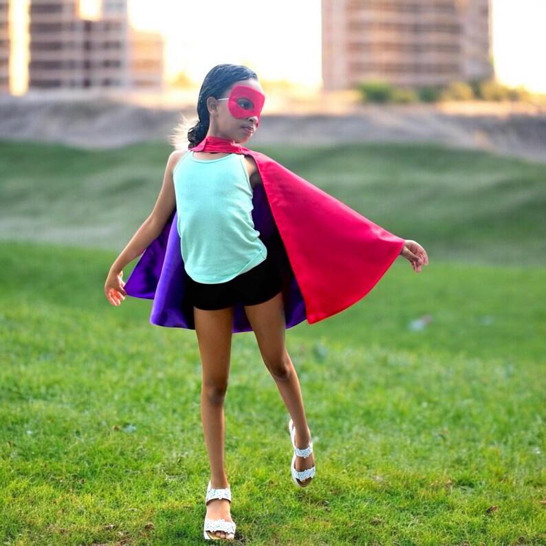 May include: A child wearing a superhero costume, including a red mask, a red and purple cape, a light blue tank top, and black shorts. The child is standing on green grass, with buildings in the background.